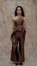Woman wearing a brown one-shoulder top and wide-leg pants with intricate detailing against a patterned backdrop.