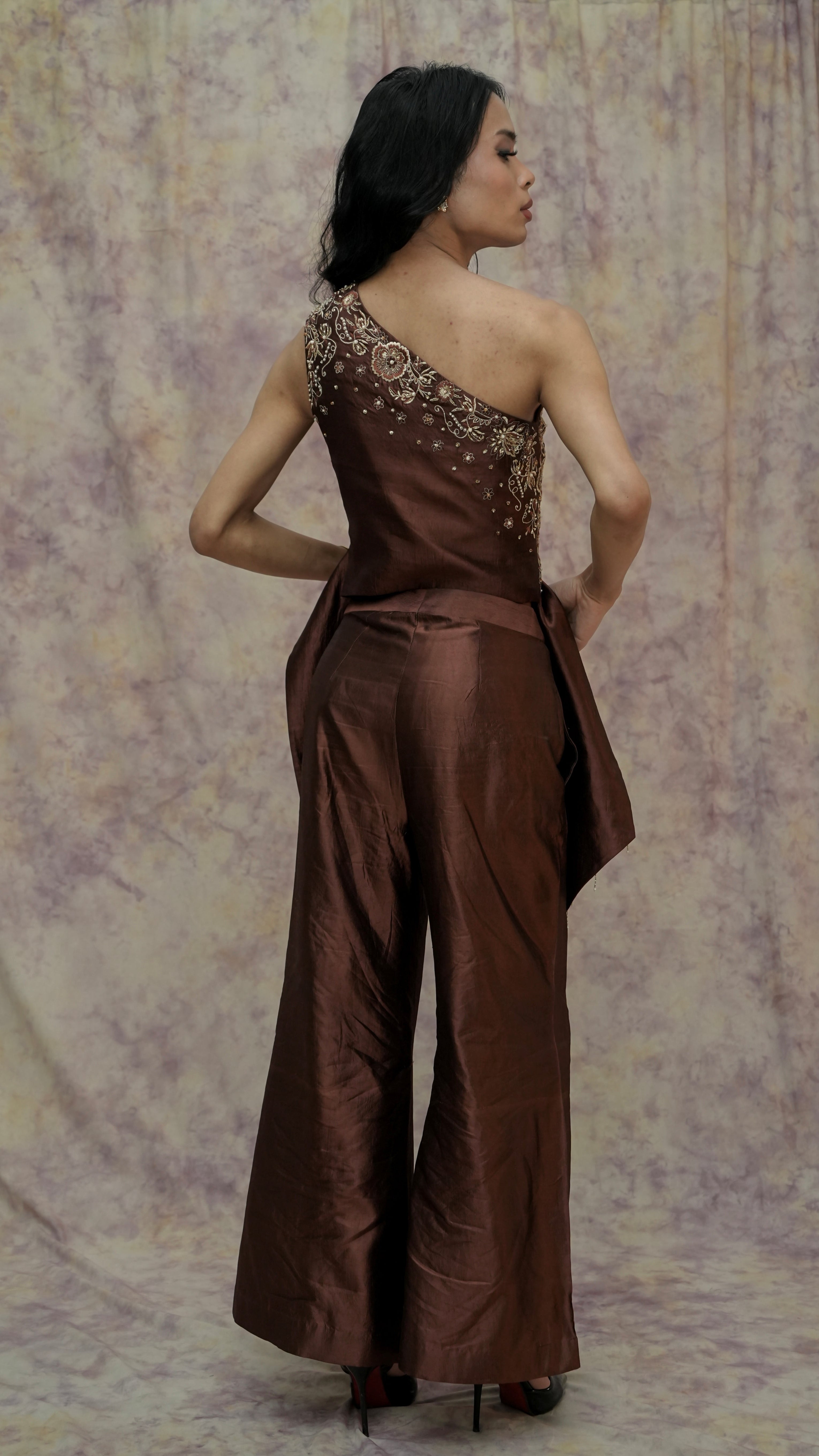 Woman wearing a brown one-shoulder jumpsuit with floral embellishments against a patterned backdrop.