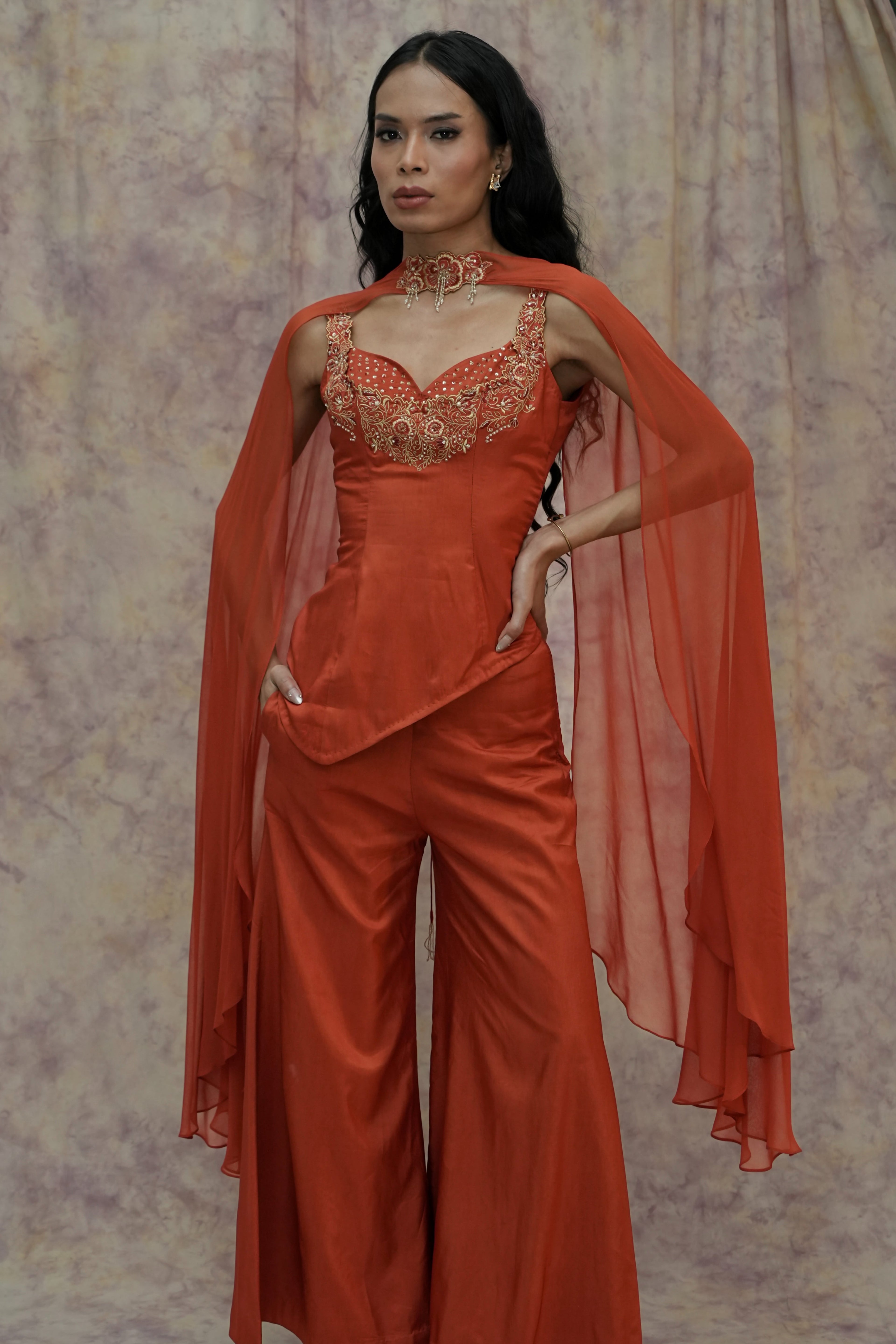 Woman wearing an orange outfit with a cape against a textured wall.