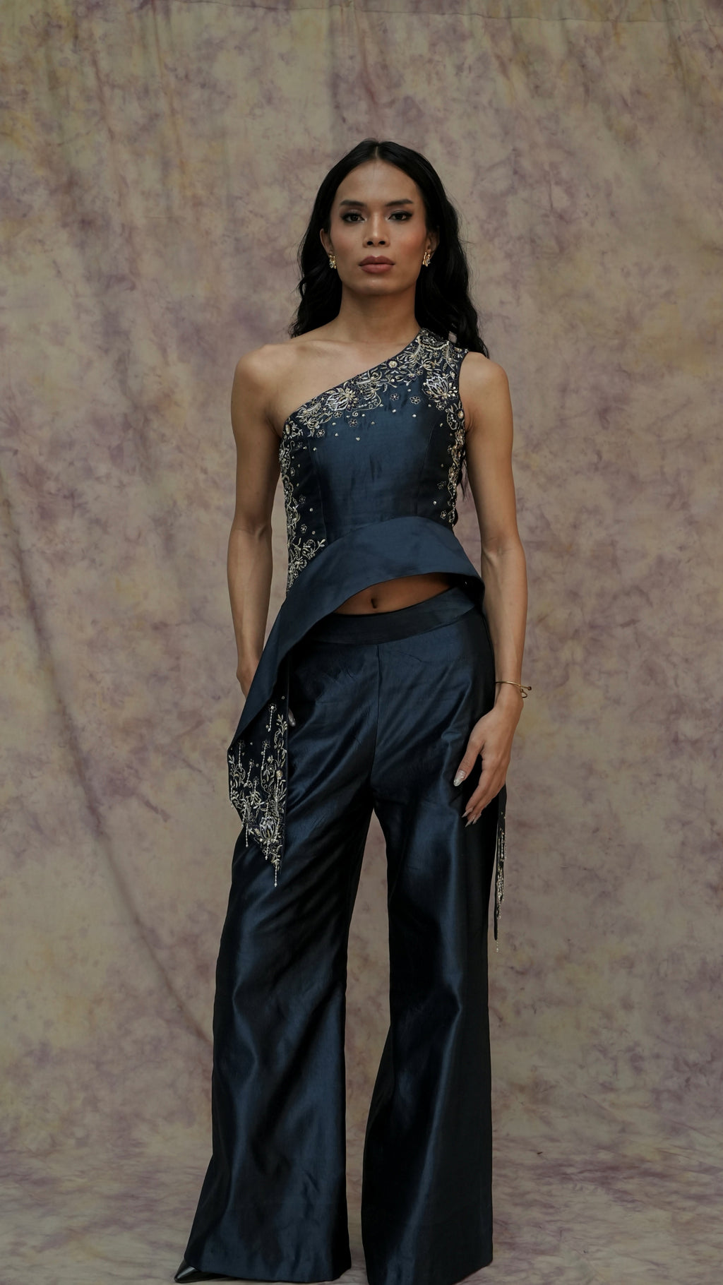 Woman wearing a dark blue one-shoulder outfit with embellishments against a textured beige background
