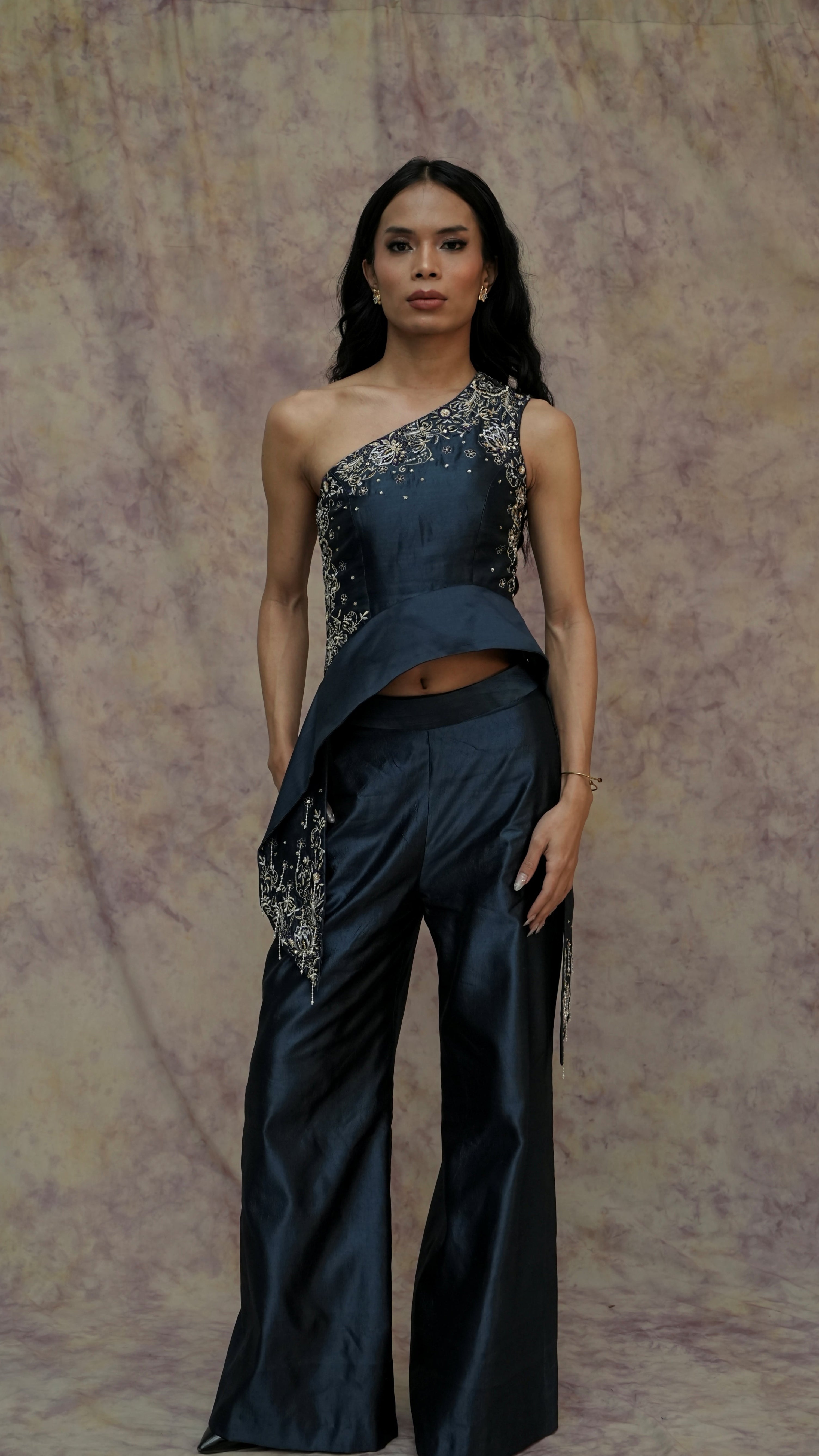 Woman wearing a dark blue one-shoulder outfit with embellishments against a textured beige background