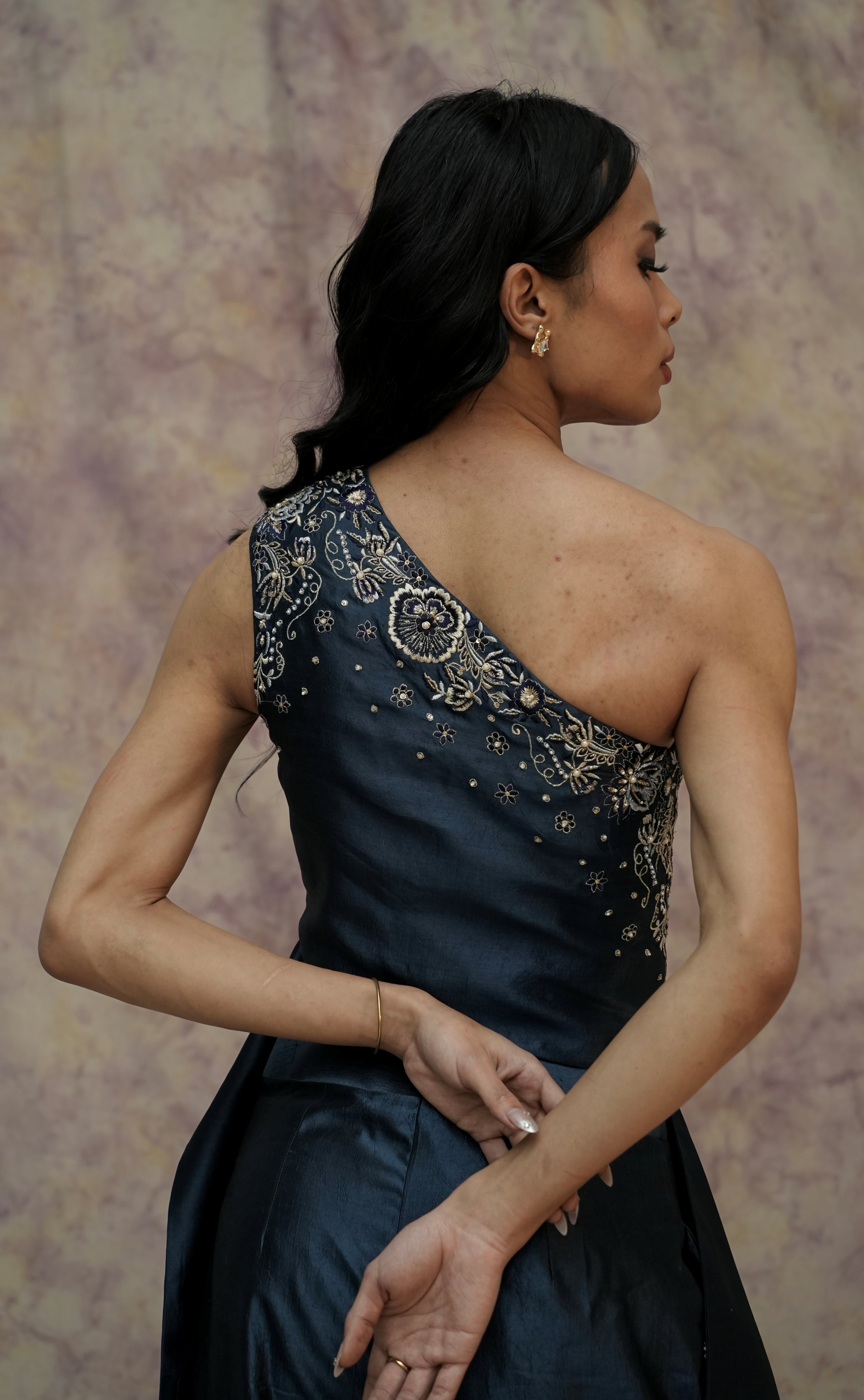 Woman wearing a dark blue one-shoulder dress with intricate gold embroidery against a textured beige wall.