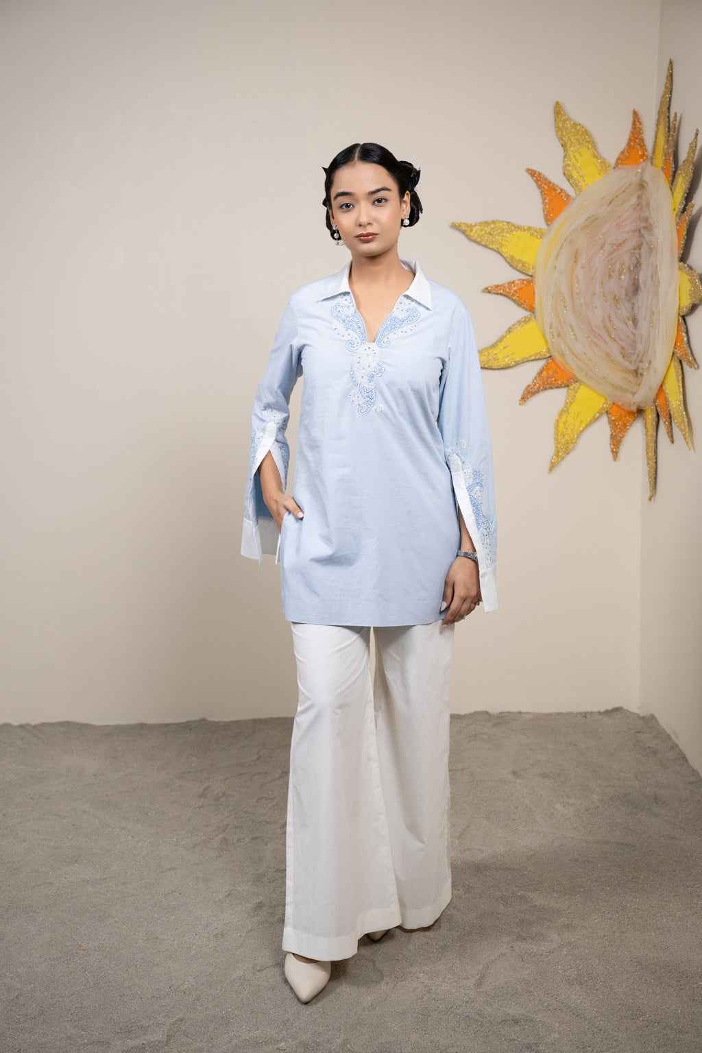Woman in a light blue shirt and white pants standing in front of a sun art piece.