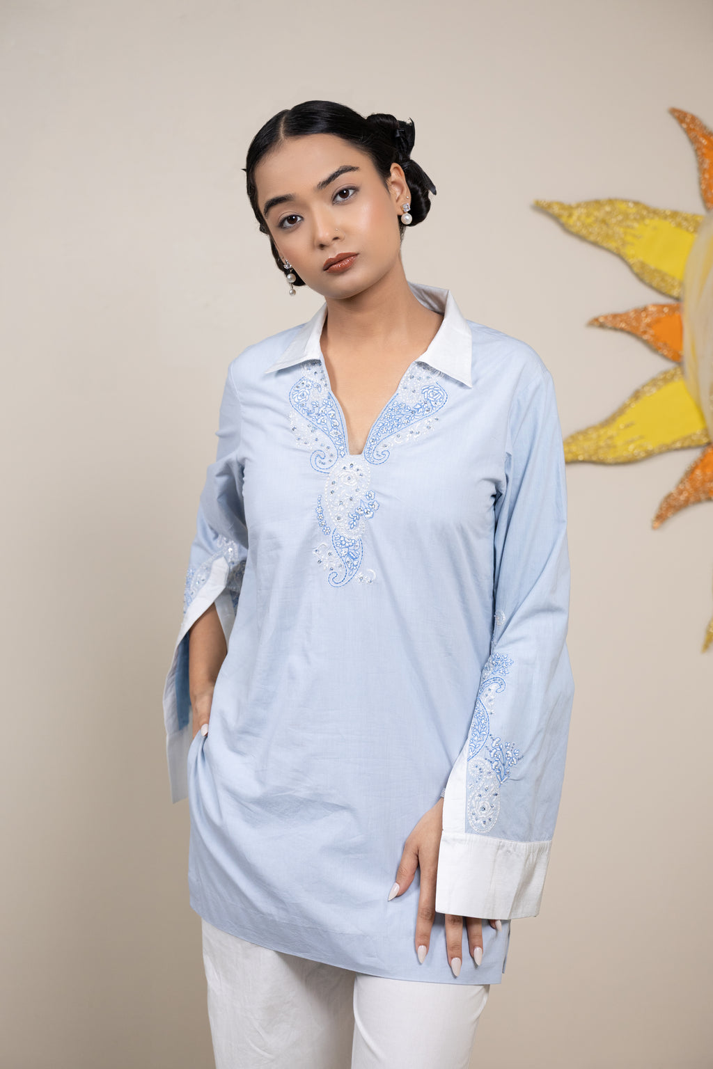 Woman wearing a light blue embroidered blouse with white pants against a beige background