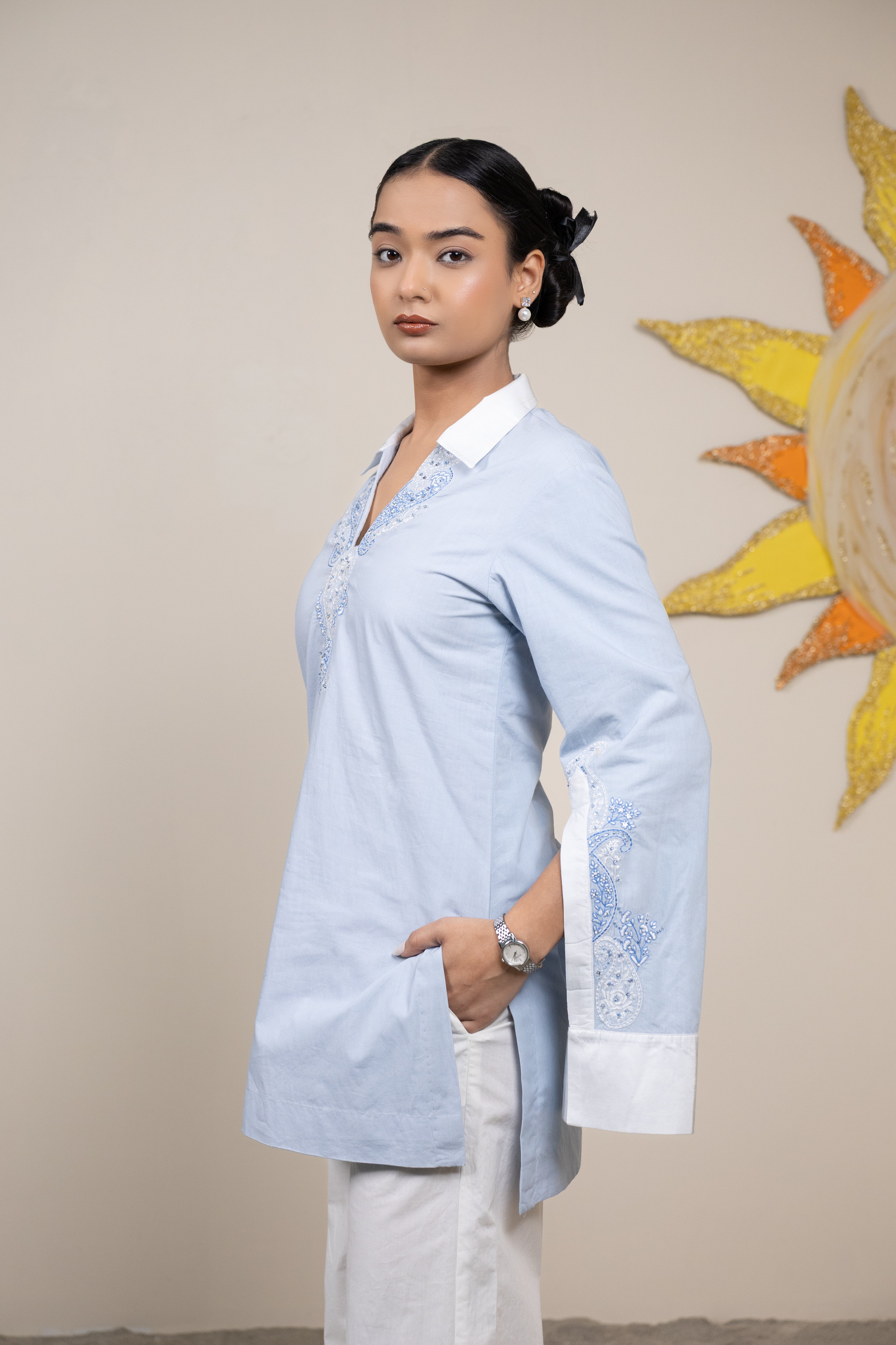 Woman wearing a light blue embroidered blouse with white pants against a beige wall with a sun decoration.
