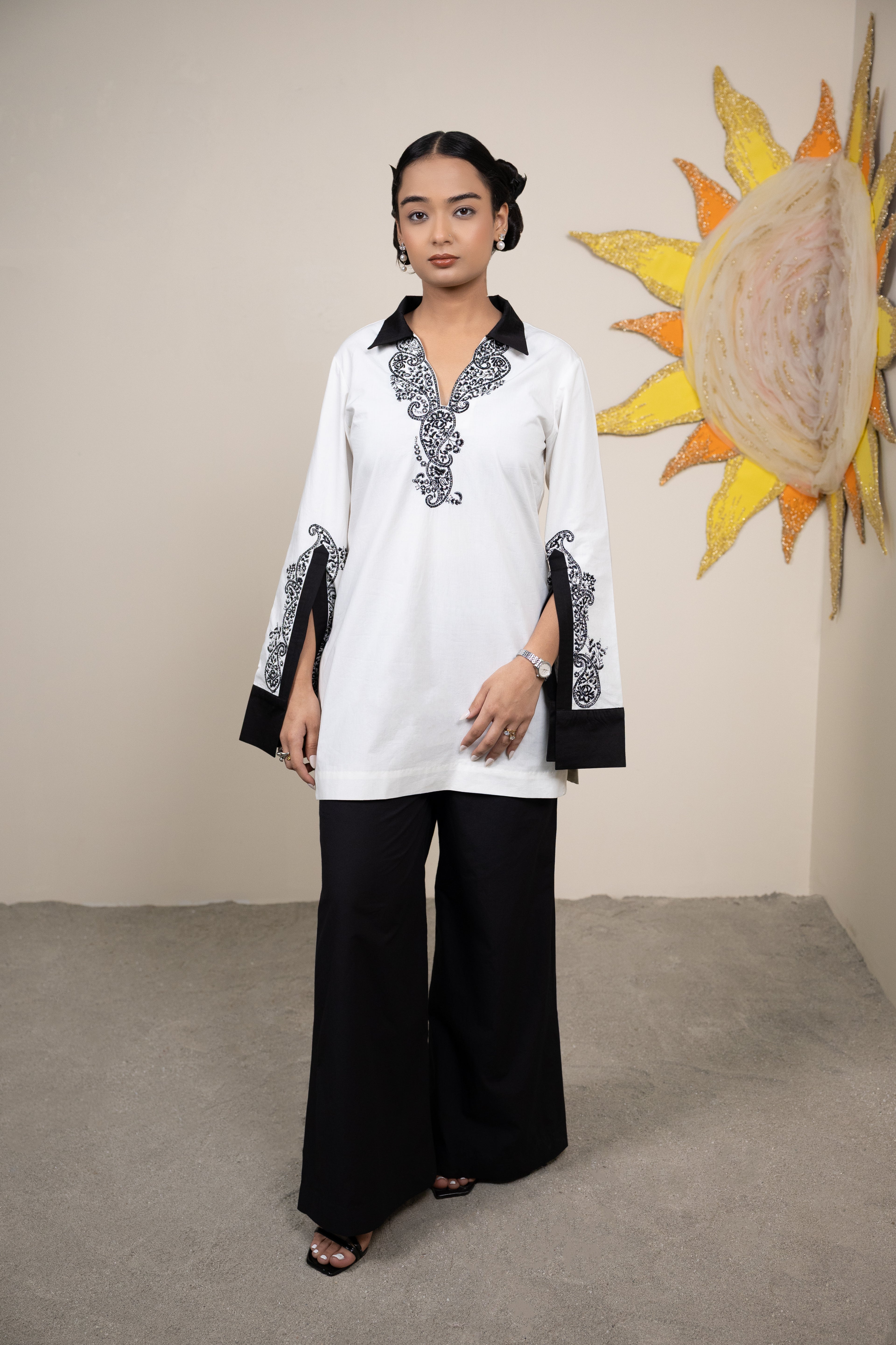 Woman wearing a white and black outfit with decorative elements, standing in front of a sunflower painting.