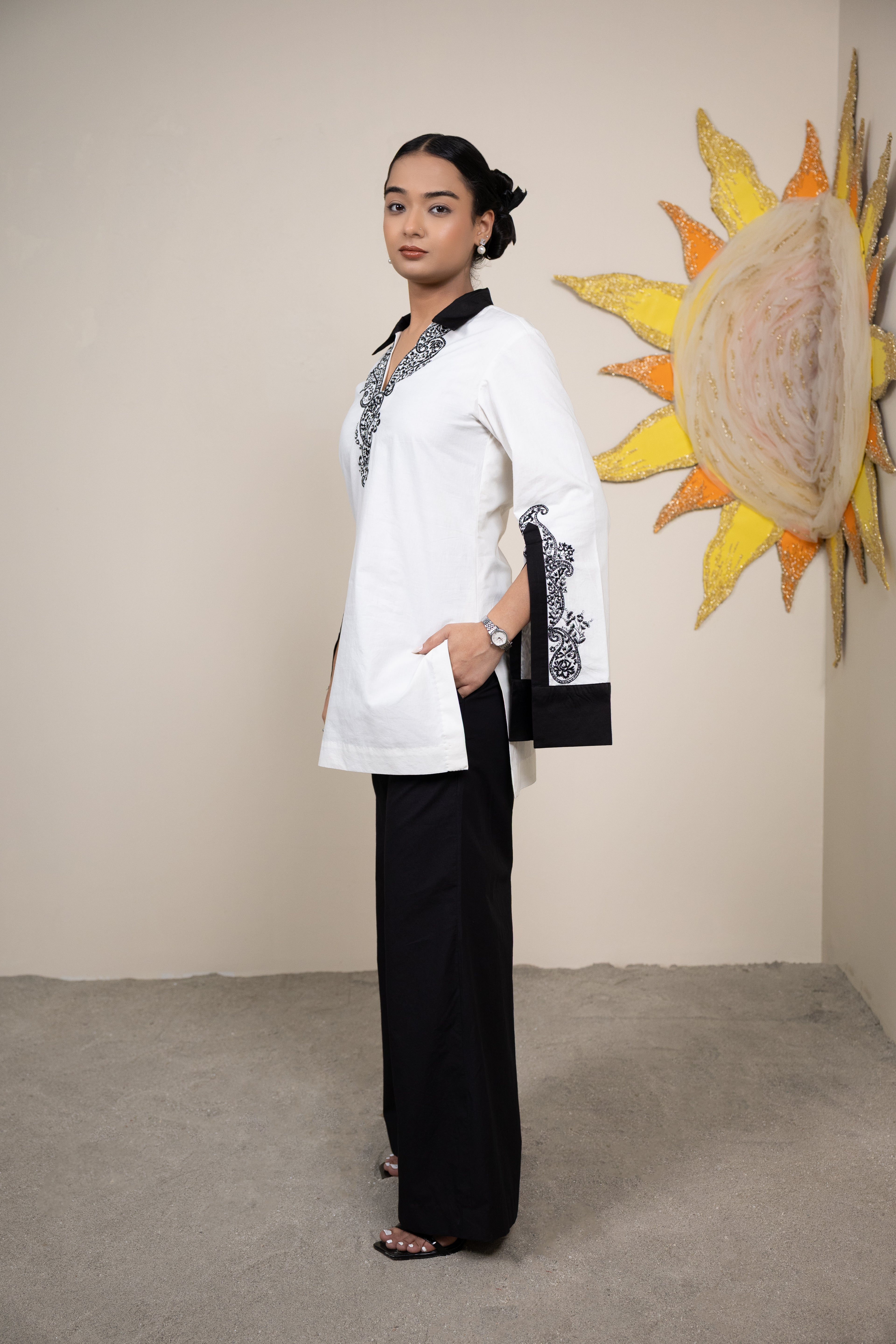 Woman in a white and black outfit standing in front of a wall with a sun and shell decoration.