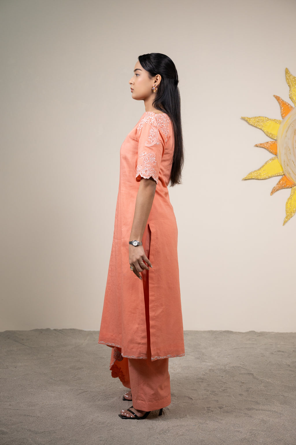 Woman in a peach kurta standing against a plain wall with a decorative sun wall art.