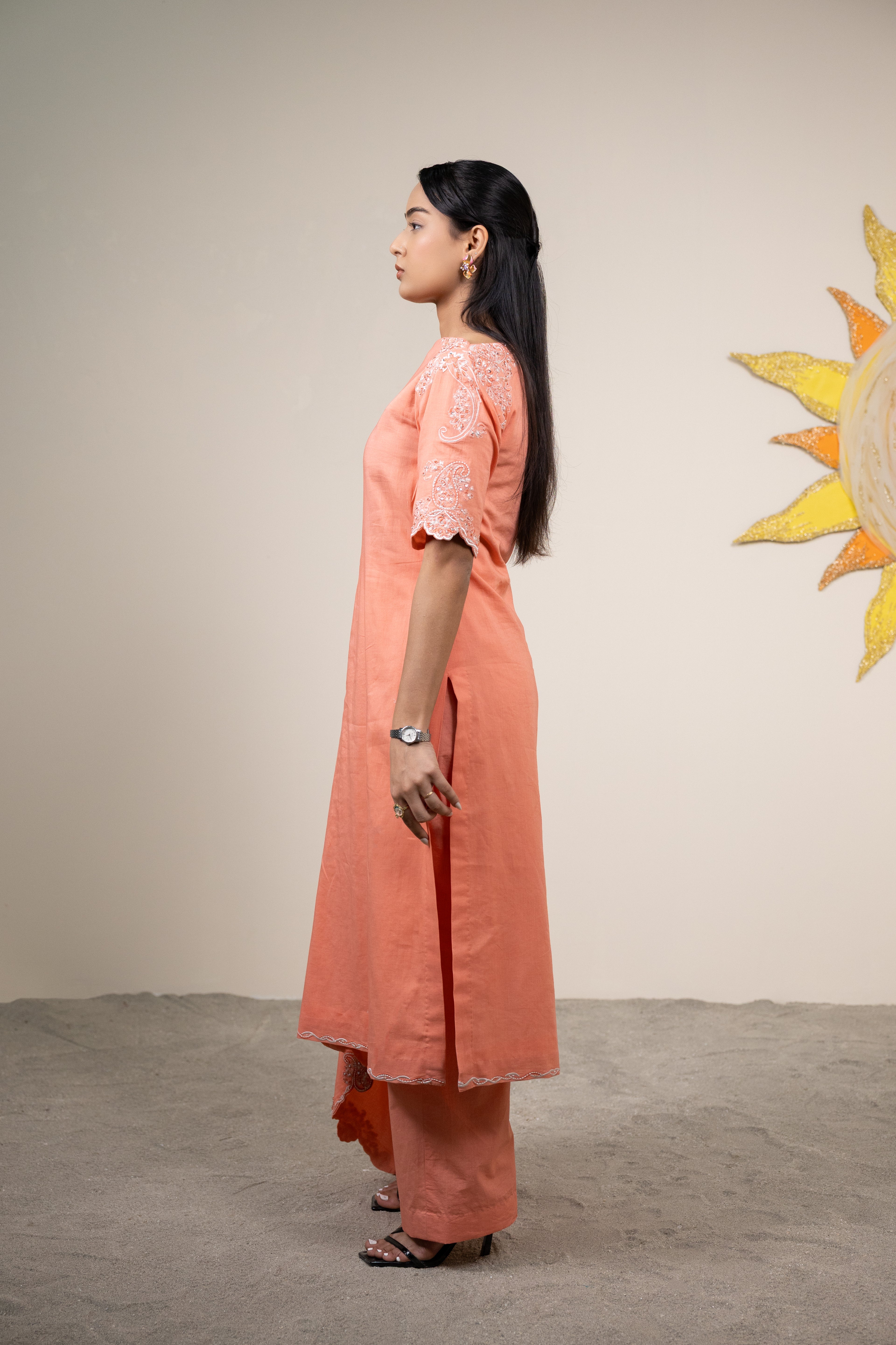 Woman in a peach kurta standing against a plain wall with a decorative sun wall art.