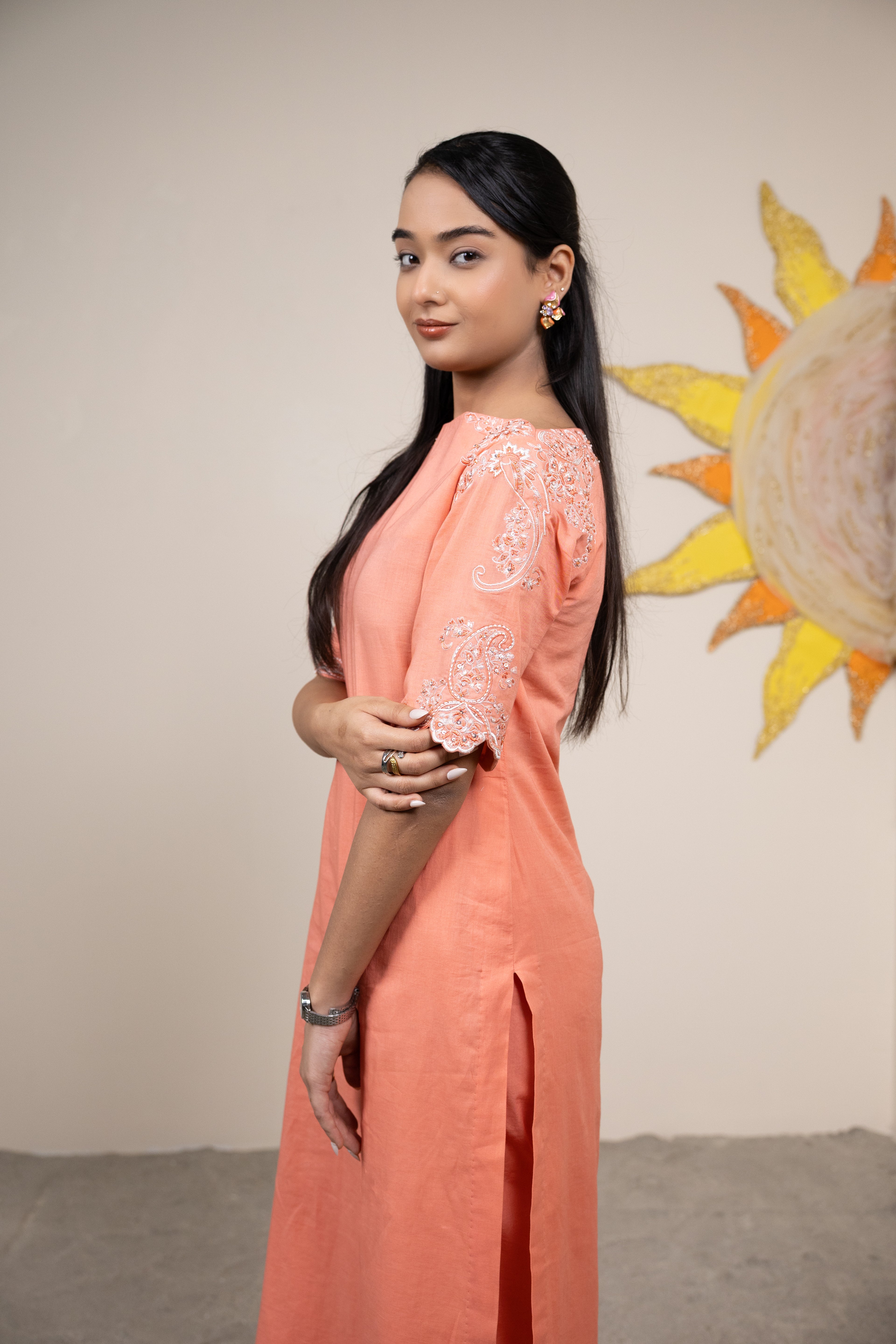 Woman in a peach outfit with floral embroidery standing in front of a sun mural.