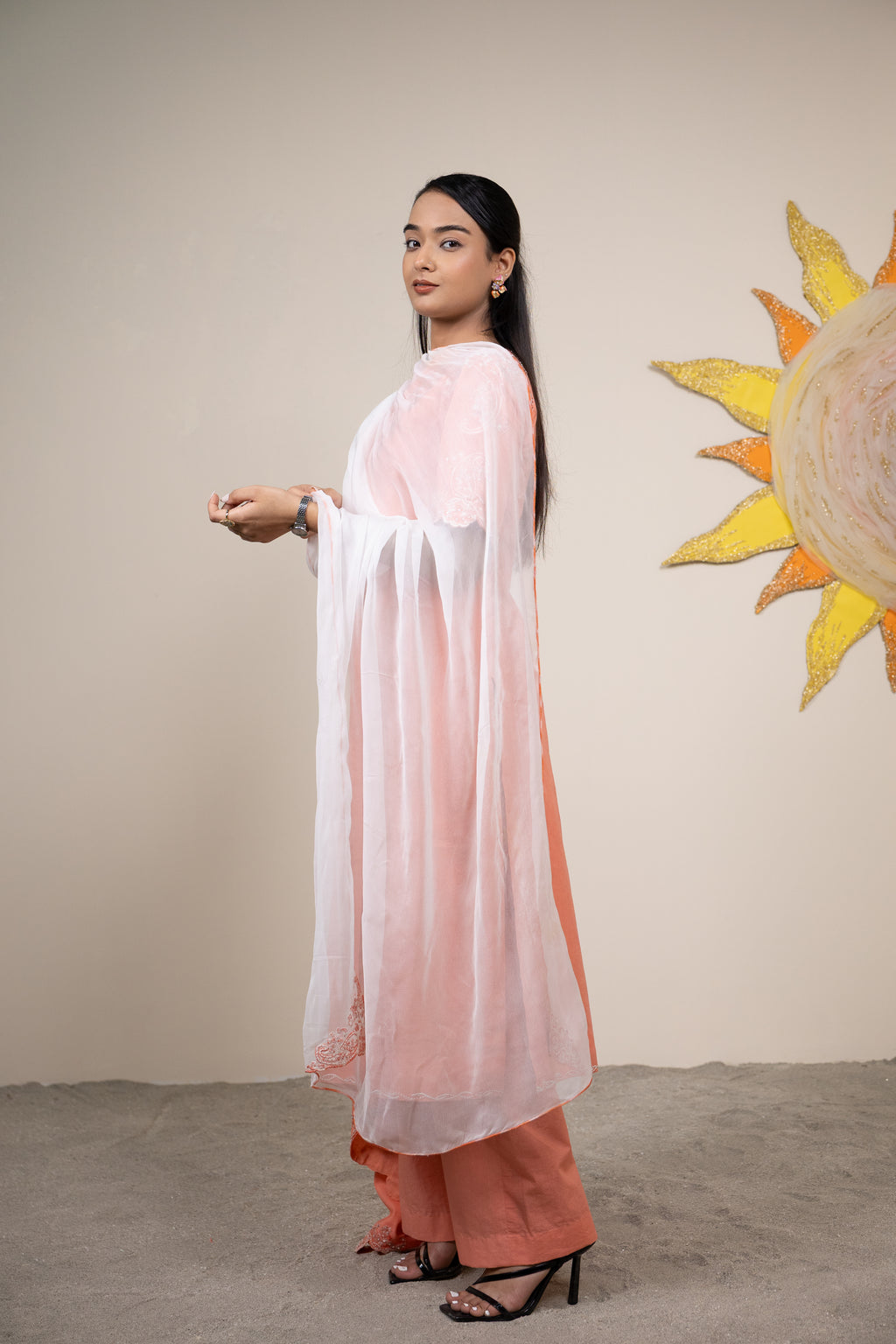 Woman in a light pink and orange outfit standing against a beige wall with a sun painting.