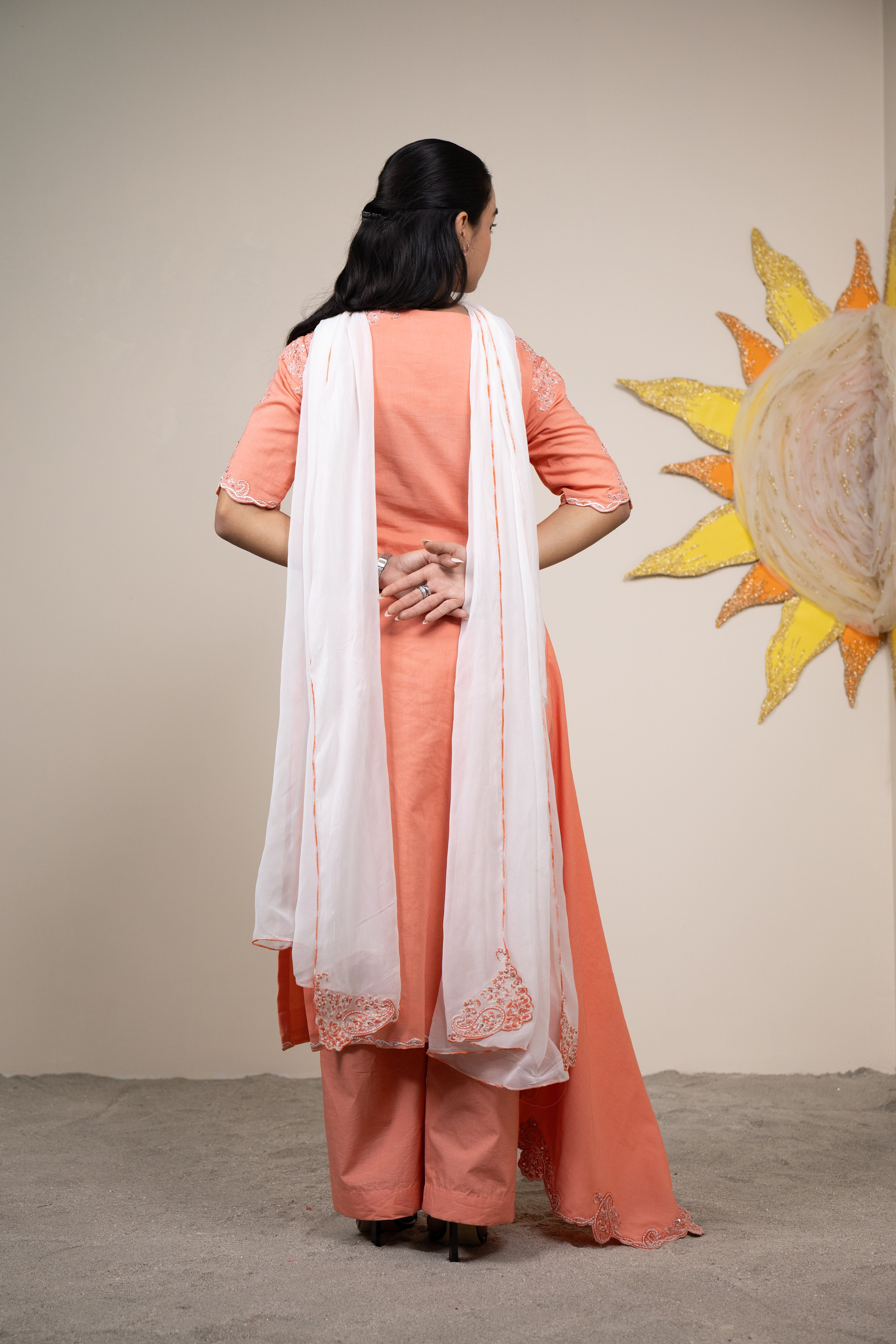 Woman in peach traditional outfit with white dupatta standing in front of a sun wall art.