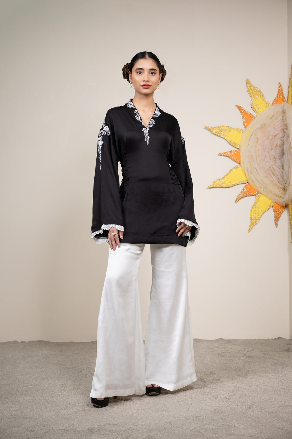 Woman wearing a black top and white pants standing in front of a decorative wall piece.