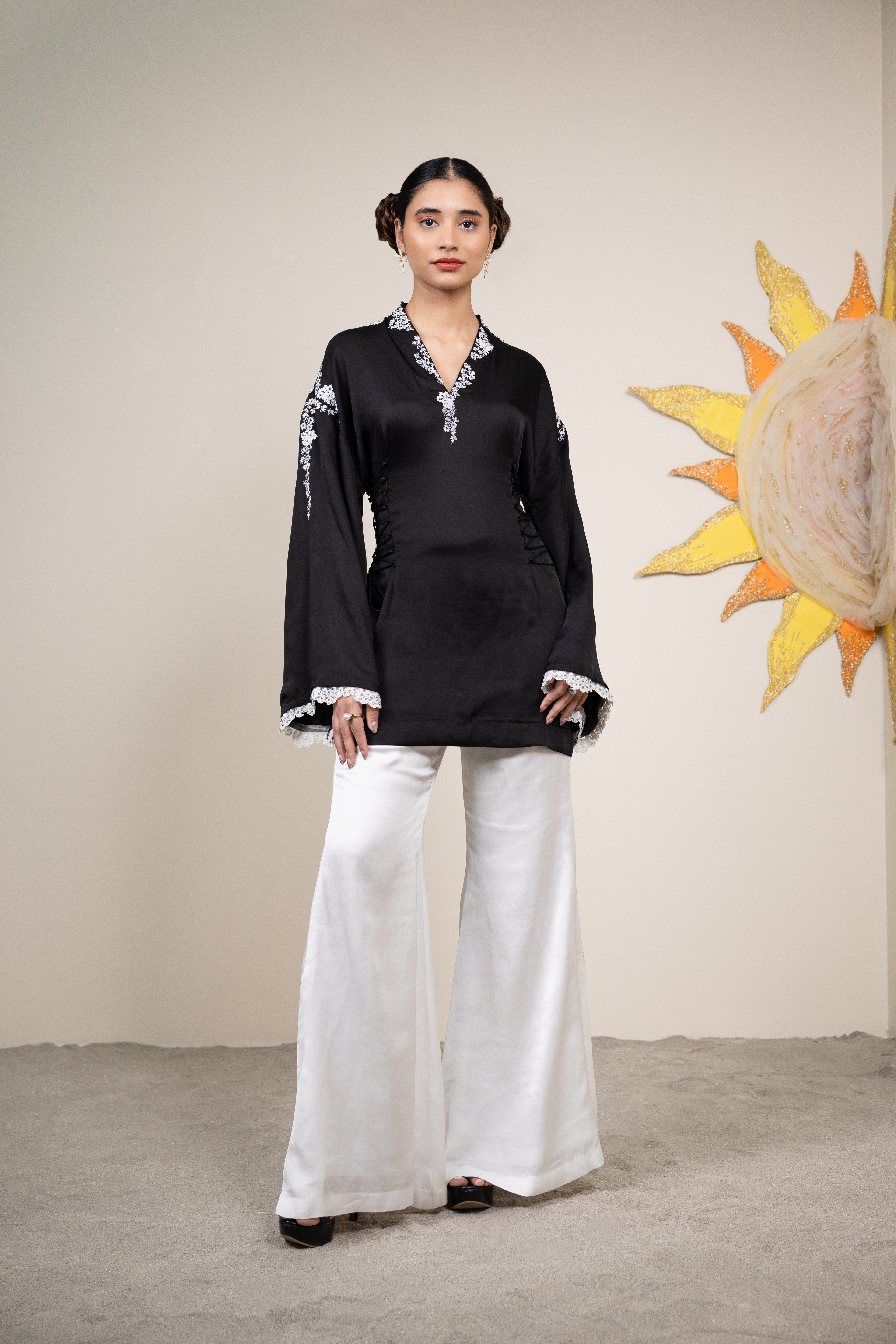 Woman wearing a black top and white pants standing in front of a decorative wall piece.