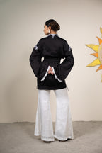 Person wearing a black traditional outfit with white patterns and wide-leg pants, standing against a beige wall with a sun decoration.