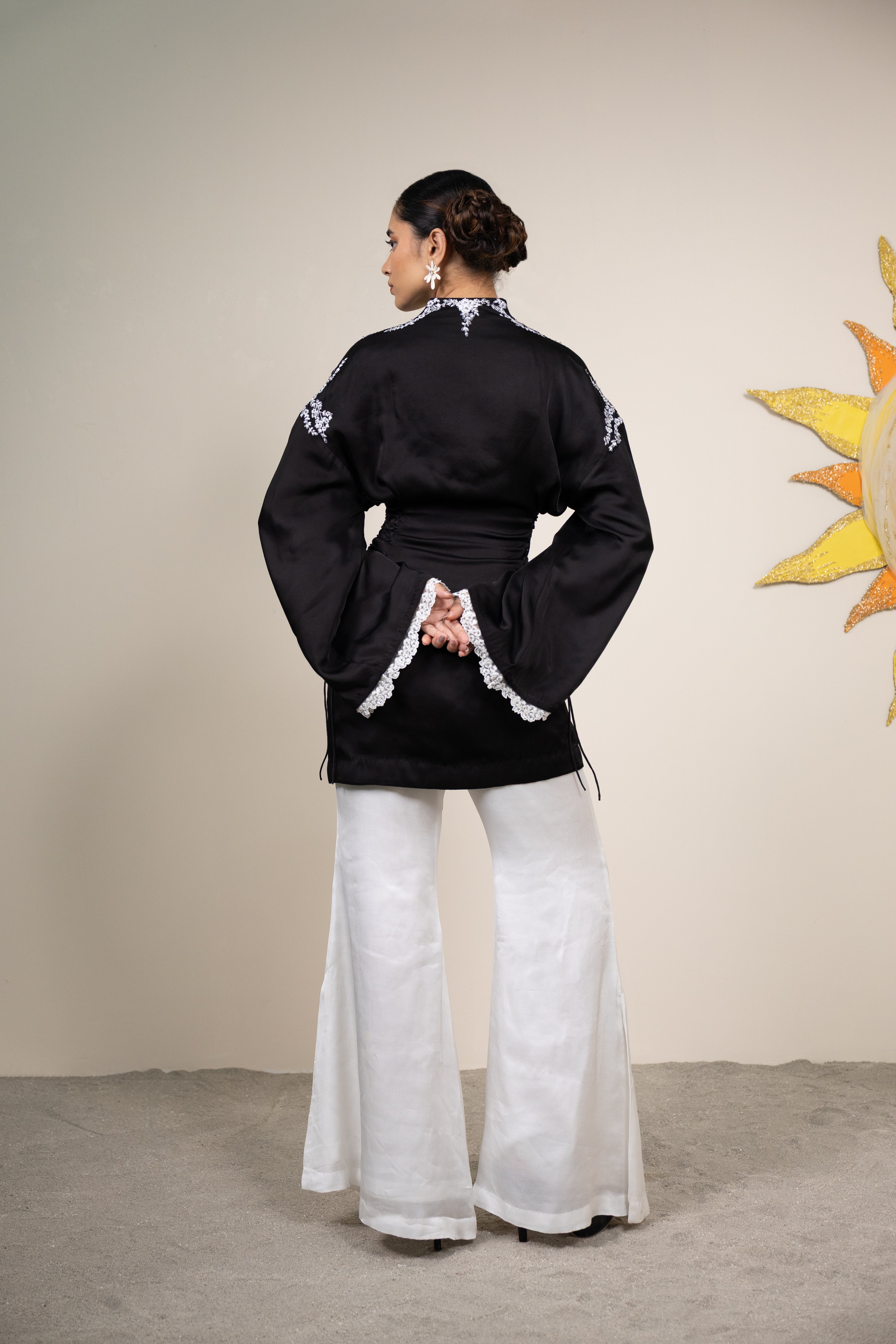 Person wearing a black traditional outfit with white patterns and wide-leg pants, standing against a beige wall with a sun decoration.
