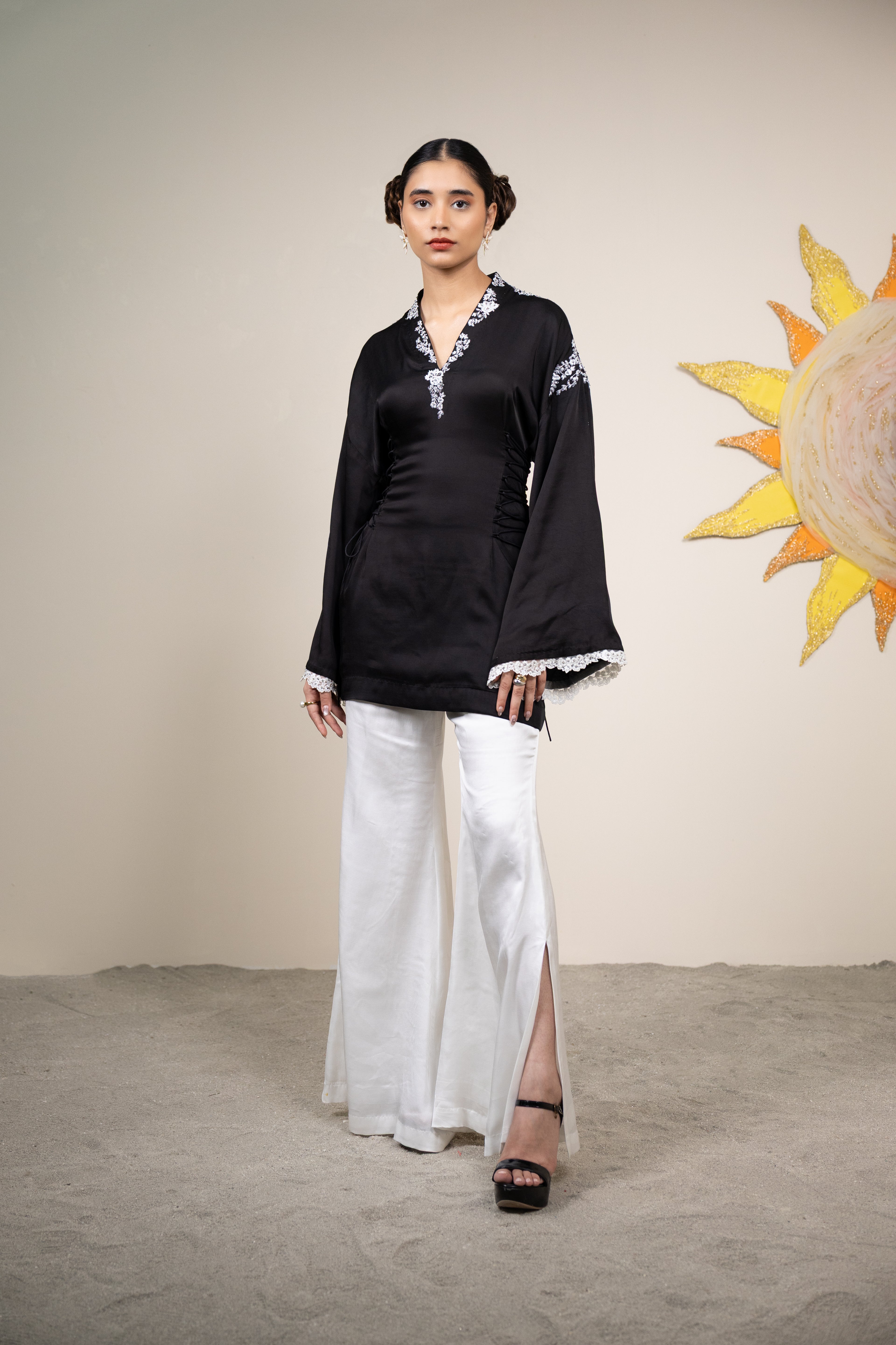Woman wearing a black top with white embroidery and white pants against a beige wall with a sun decoration.