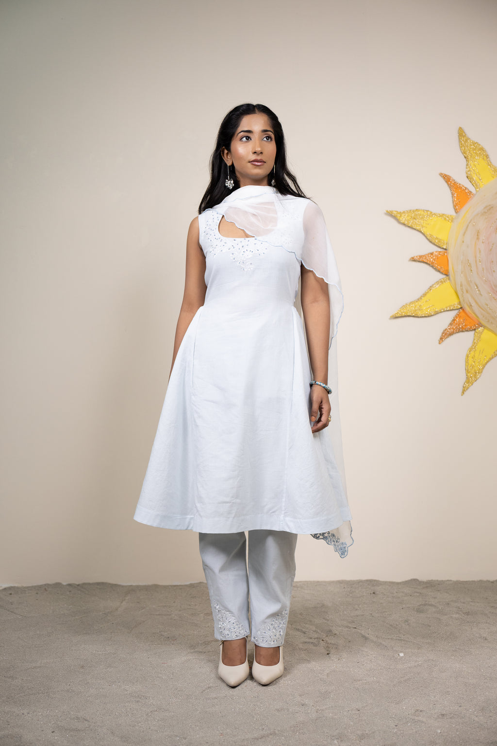 Woman wearing a white dress with a cutout design, standing against a beige wall with a sun painting.