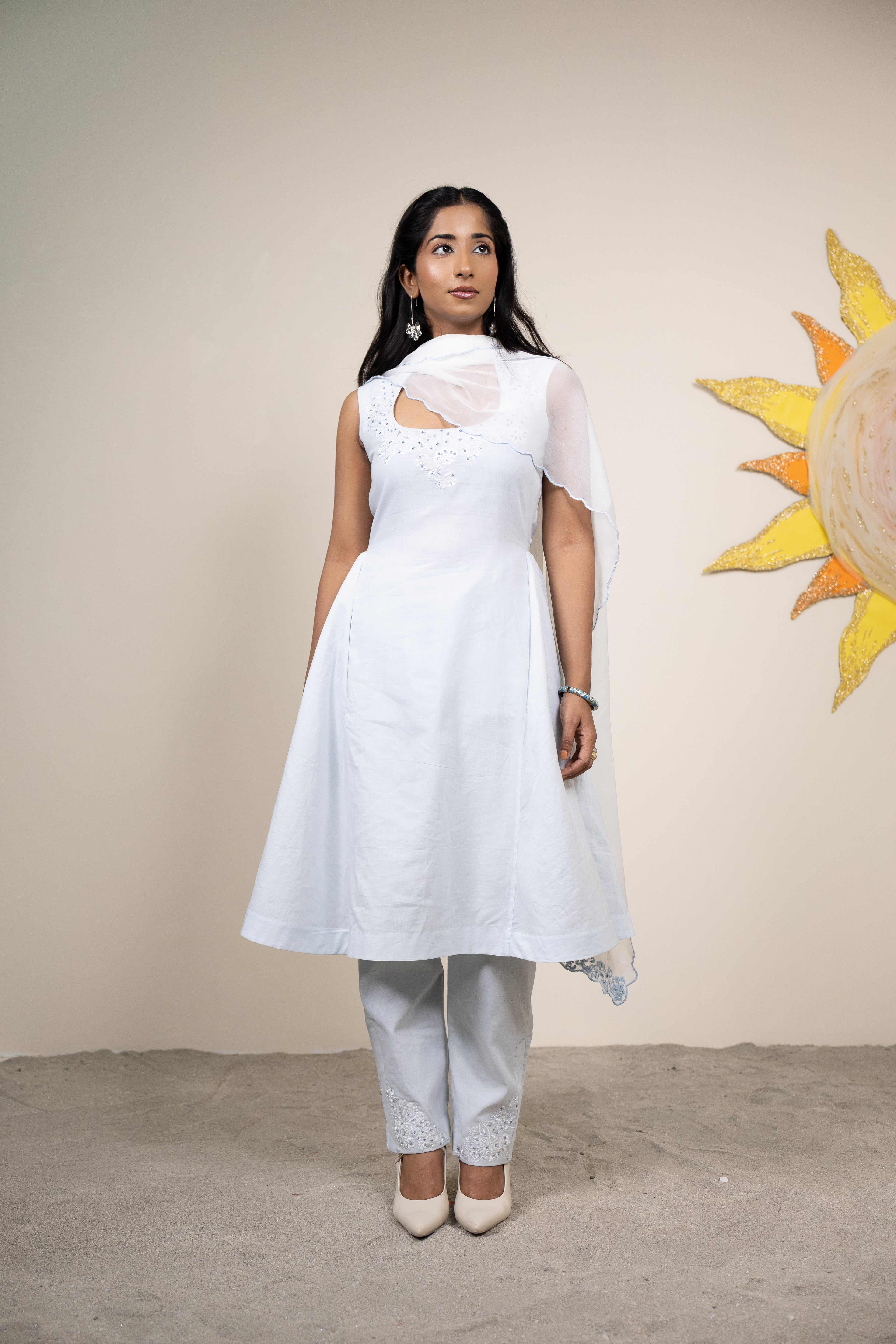 Woman wearing a white dress with a cutout design, standing against a beige wall with a sun painting.