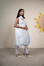 Woman in a white traditional outfit standing against a beige wall with a sun design.