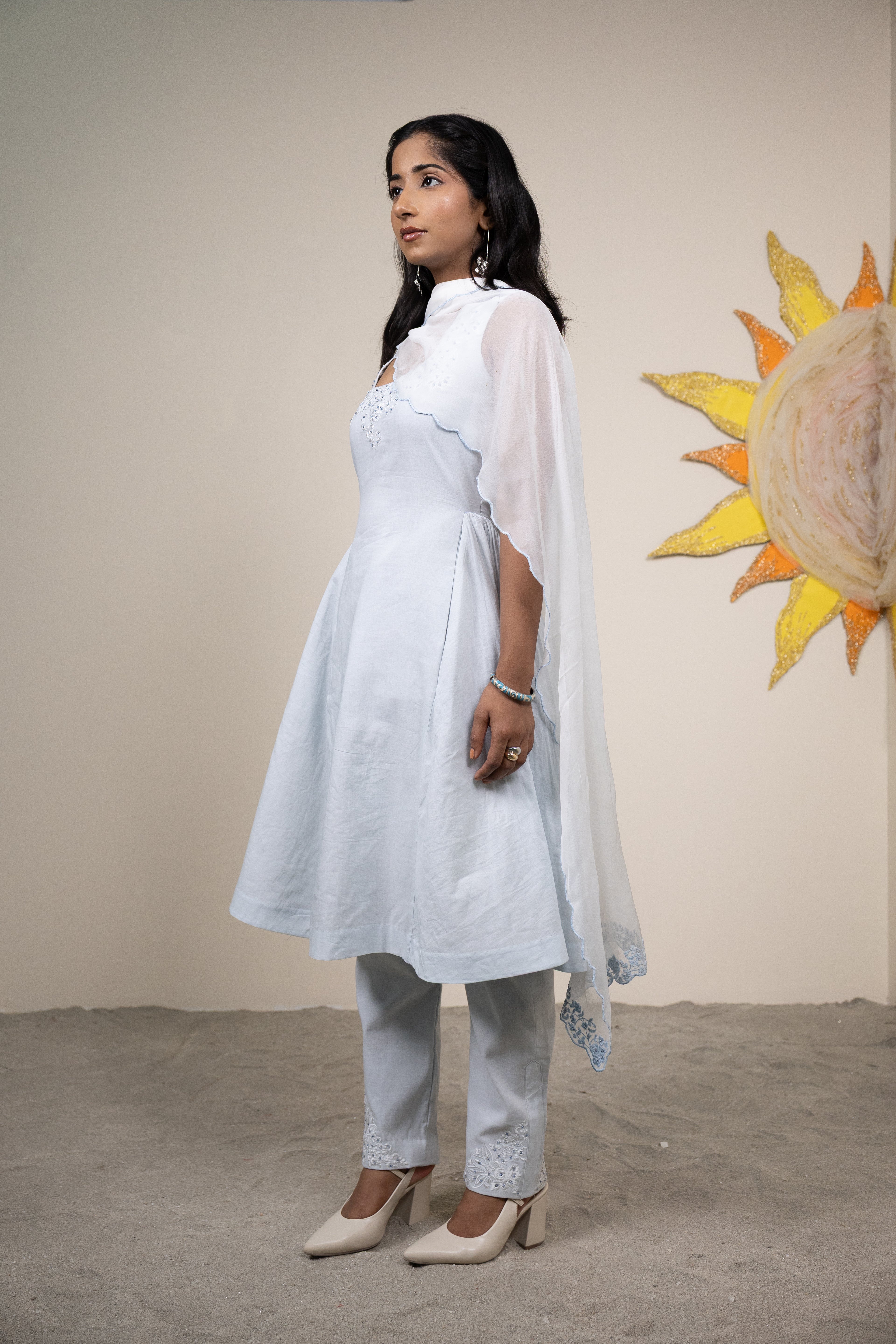 Woman in a white outfit standing in front of a beige wall with a sun painting.